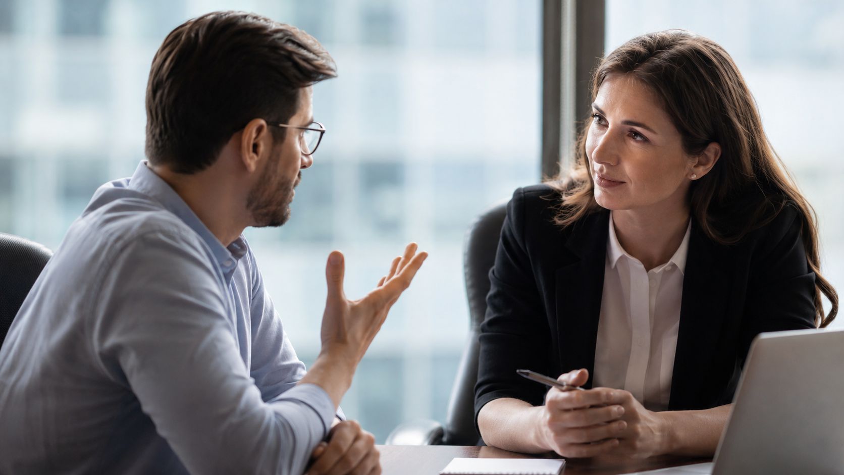 Two colleagues seated at a meeting table in a focused conversation, one gesturing while the other listens.